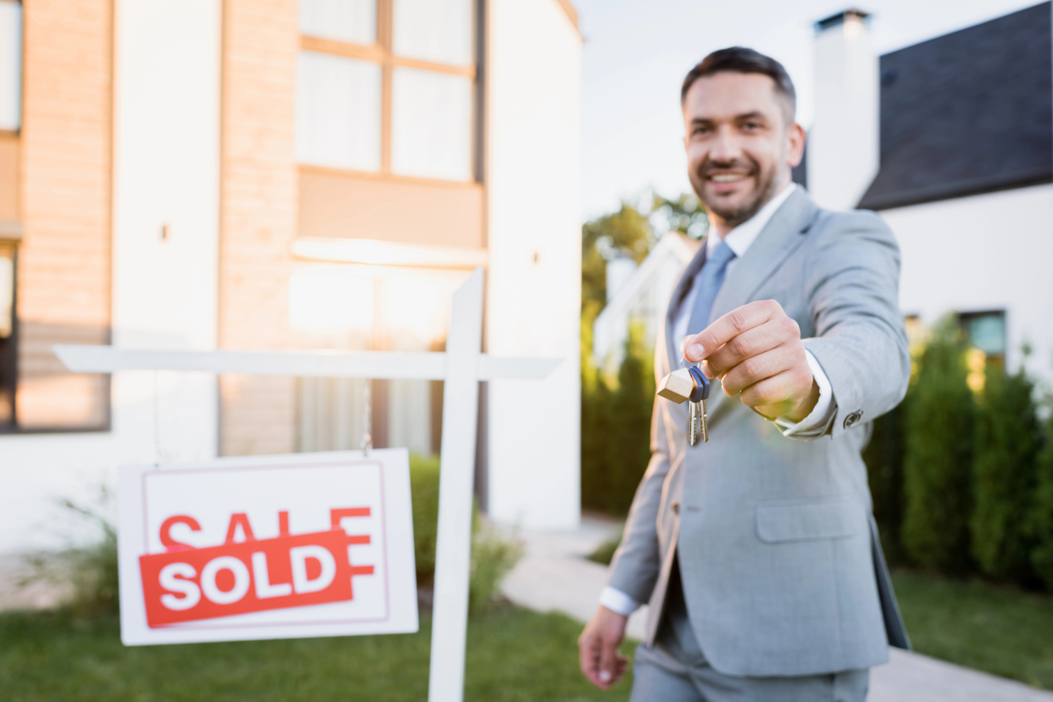 Smiling broker looking at camera while showing keys near sign with sold lettering on blurred background